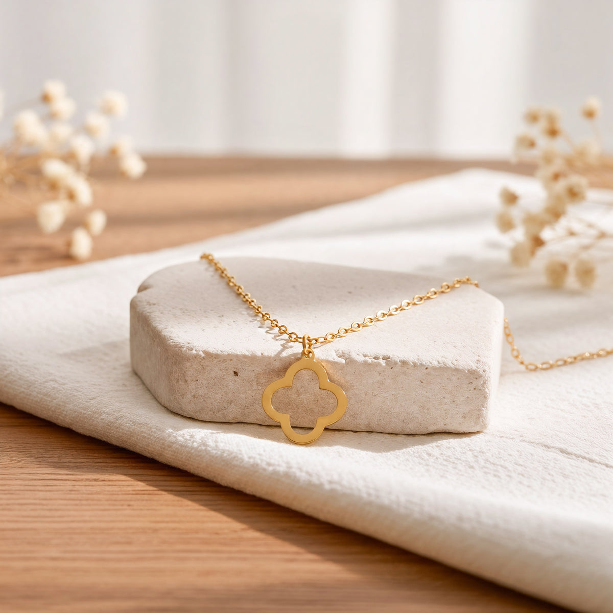 Kleeblatt-Halskette aus Edelstahl 18 K vergoldet, liegt auf hellem Stoff und Steinpodest mit Trockenblumen im Hintergrund – Thaleya Halskette | Mercy’s Jewelry