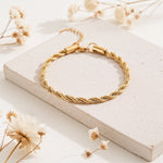 Gedrehtes Seilarmband aus Edelstahl 18 K vergoldet, elegant auf heller Steinplatte mit Trockenblumen arrangiert – Tharien Armband | Mercy’s Jewelry