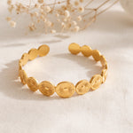 Armreif aus Edelstahl 18 K vergoldet mit gravierten Sonnensymbolen, liegend auf weißem Stoff mit Trockenblumen im Hintergrund – Aurema Armreif | Mercy’s Jewelry