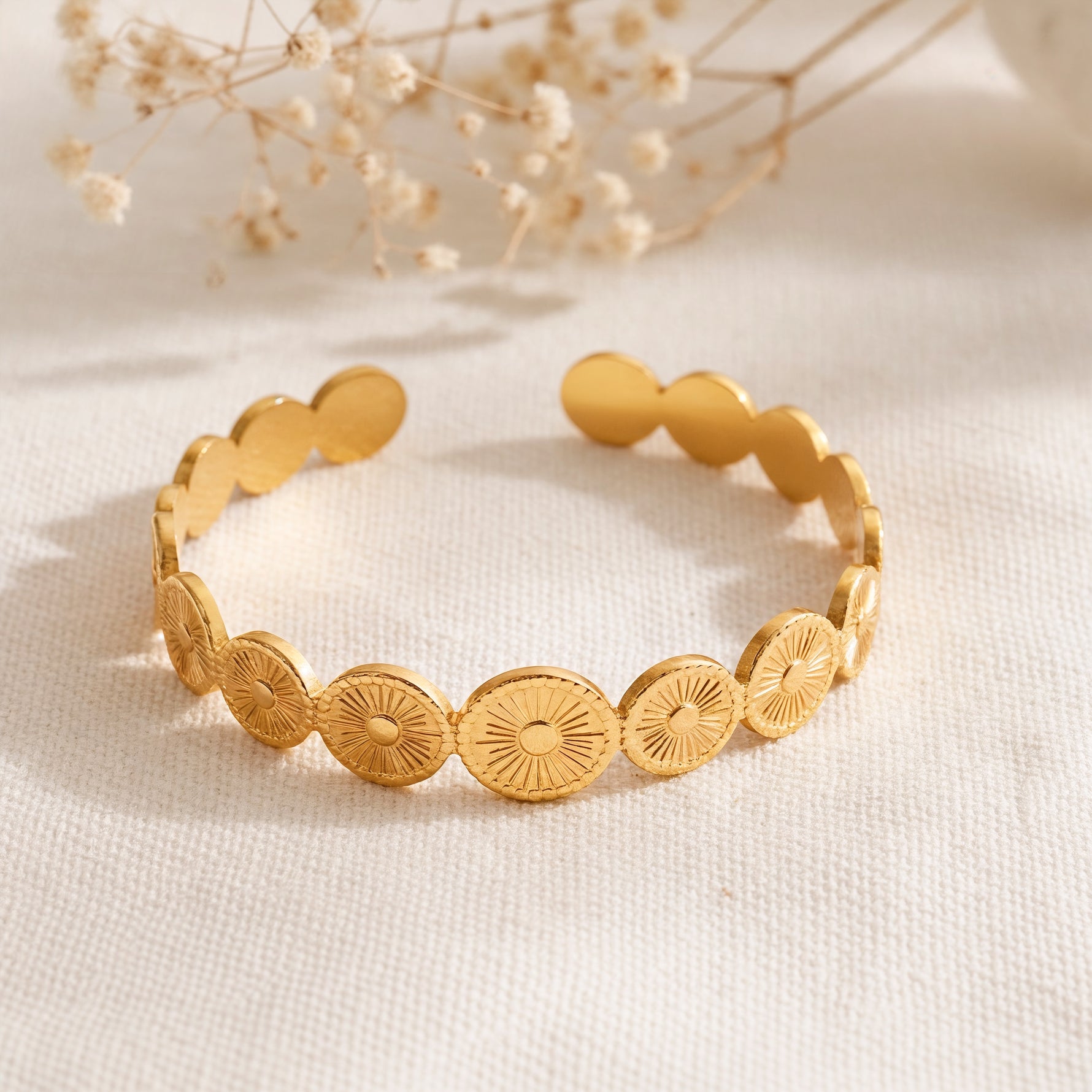 Armreif aus Edelstahl 18 K vergoldet mit gravierten Sonnensymbolen, liegend auf weißem Stoff mit Trockenblumen im Hintergrund – Aurema Armreif | Mercy’s Jewelry