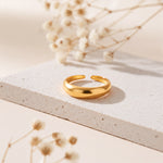 Bandring aus Edelstahl 18 K vergoldet, schlichtes gewölbtes Design auf heller Steinplattform mit Trockenblumen im Hintergrund – Aralyn Ring | Mercy’s Jewelry