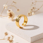 Breiter Bandring aus Edelstahl 18 K vergoldet, glänzendes gewölbtes Design auf heller Steinplattform mit Trockenblumen im Hintergrund – Orisha Ring | Mercy’s Jewelry