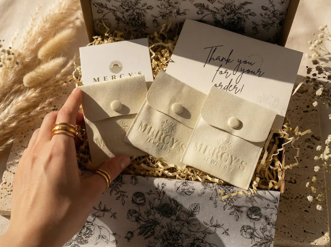 Das MERCY'S Ritual von bymercy.de: Blick in die edle Schmuck-Geschenkbox mit cremefarbenen Beuteln und Dankeskarte auf Travertinstein mit Trockenblumen.