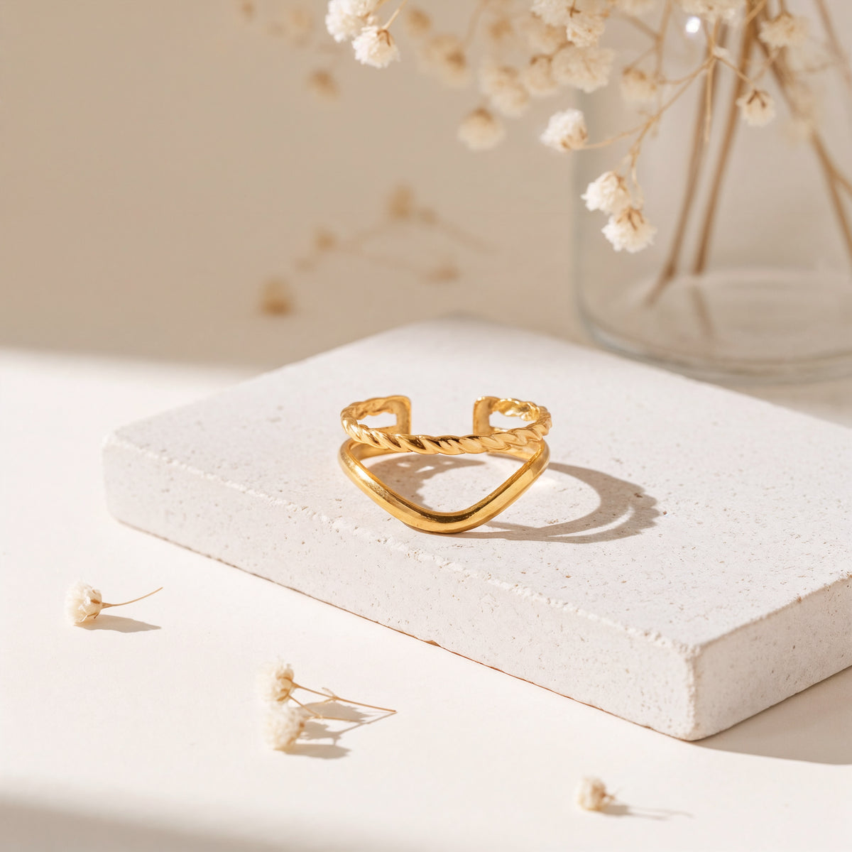 Chevron-Ring aus Edelstahl 18 K vergoldet, offen und verstellbar, mit gedrehter Struktur auf heller Steinplattform und Trockenblumen im Hintergrund – Soriel Ring | Mercy’s Jewelry