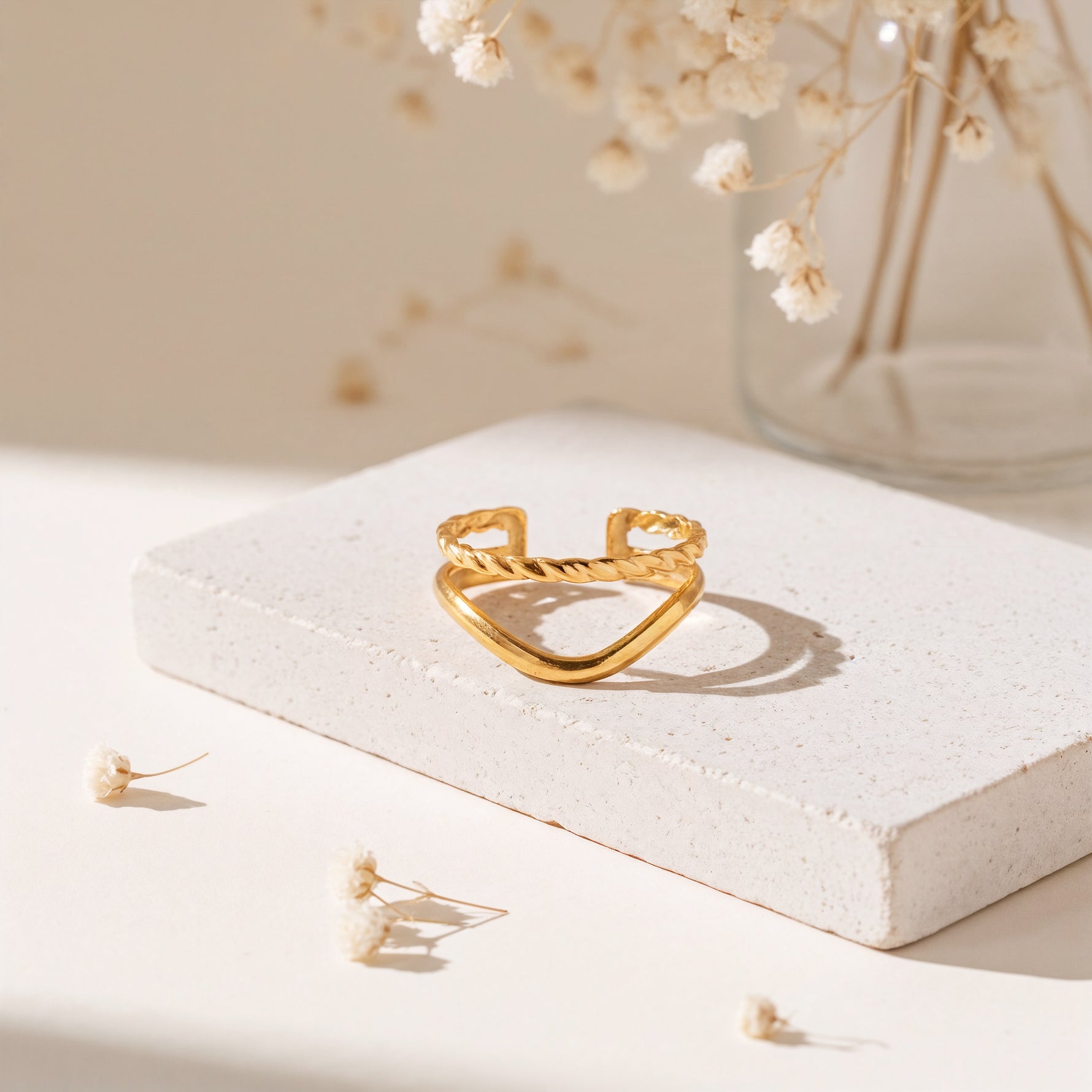 Chevron-Ring aus Edelstahl 18 K vergoldet, offen und verstellbar, mit gedrehter Struktur auf heller Steinplattform und Trockenblumen im Hintergrund – Soriel Ring | Mercy’s Jewelry