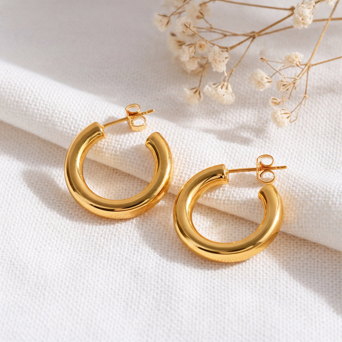 Klassische Creolen mit abgerundetem Design aus Edelstahl 18 K vergoldet, liegen auf hellem Stoff mit Trockenblumen im Hintergrund im weichen Sonnenlicht – Caelyn Creolen | Mercy’s Jewelry