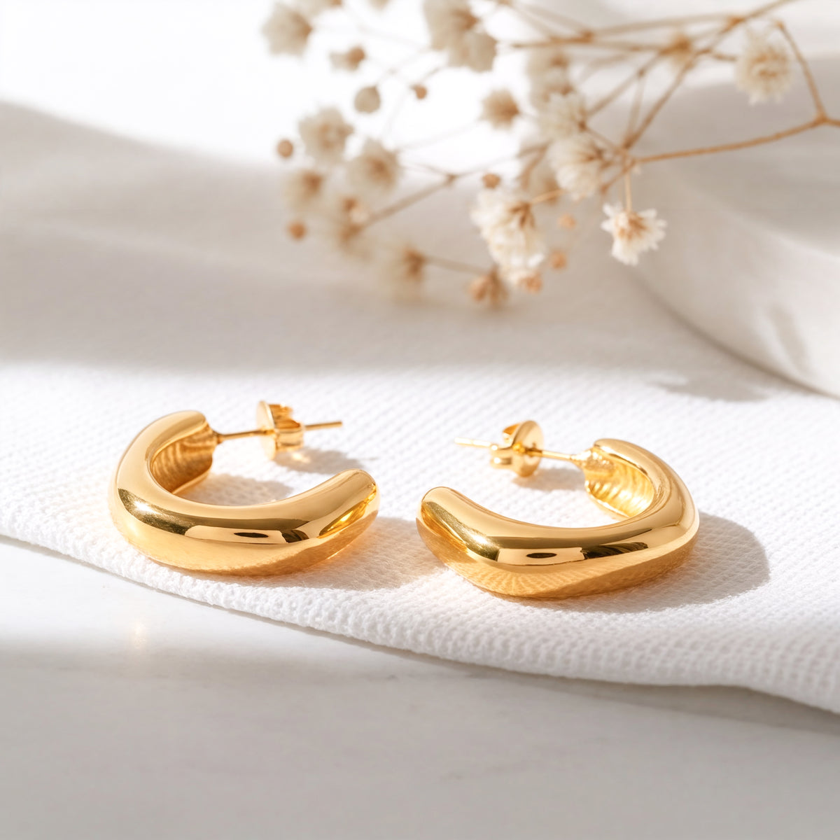 Schlichte Creolen mit abgerundetem Design aus Edelstahl 18 K vergoldet, liegen auf hellem Stoff mit Trockenblumen im Hintergrund im sanften Tageslicht – Velun Creolen | Mercy’s Jewelry