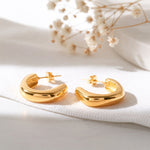 Schlichte Creolen mit abgerundetem Design aus Edelstahl 18 K vergoldet, liegen auf hellem Stoff mit Trockenblumen im Hintergrund im sanften Tageslicht – Velun Creolen | Mercy’s Jewelry