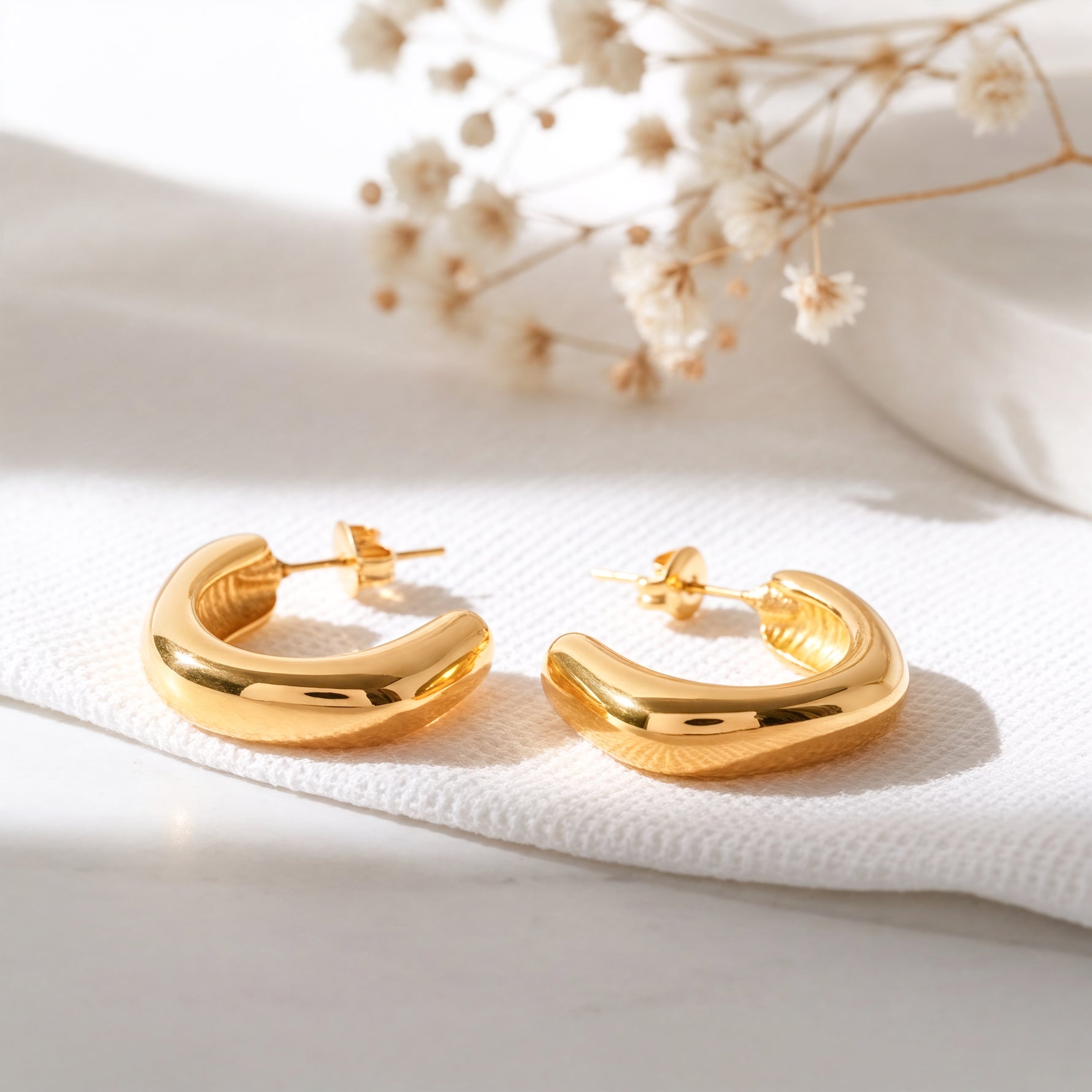 Schlichte Creolen mit abgerundetem Design aus Edelstahl 18 K vergoldet, liegen auf hellem Stoff mit Trockenblumen im Hintergrund im sanften Tageslicht – Velun Creolen | Mercy’s Jewelry