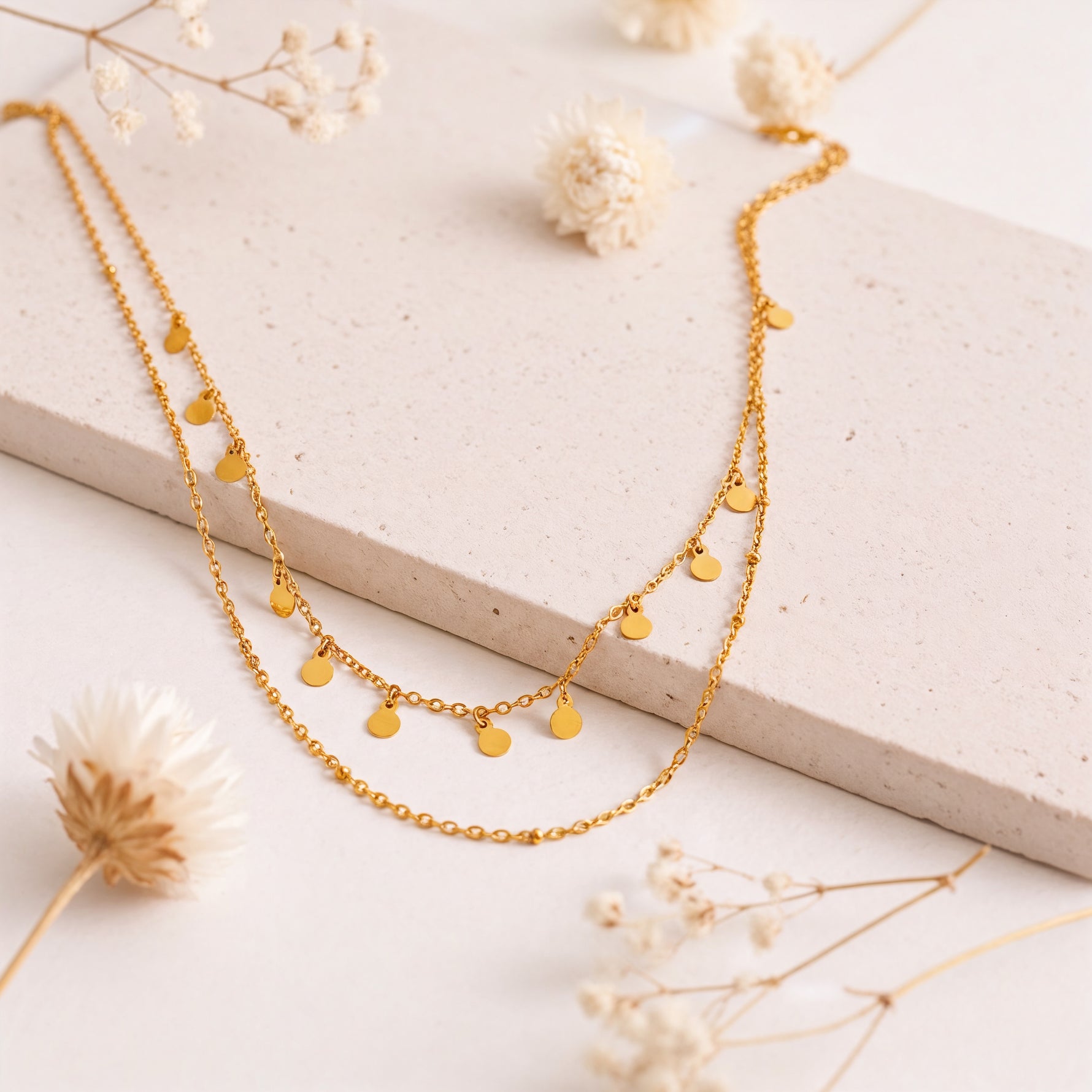 Doppellagige Halskette mit kleinen Plättchen aus Edelstahl 18 K vergoldet, liegt auf heller Steinplatte mit Trockenblumen – Naia Layers Kette | Mercy’s Jewelry