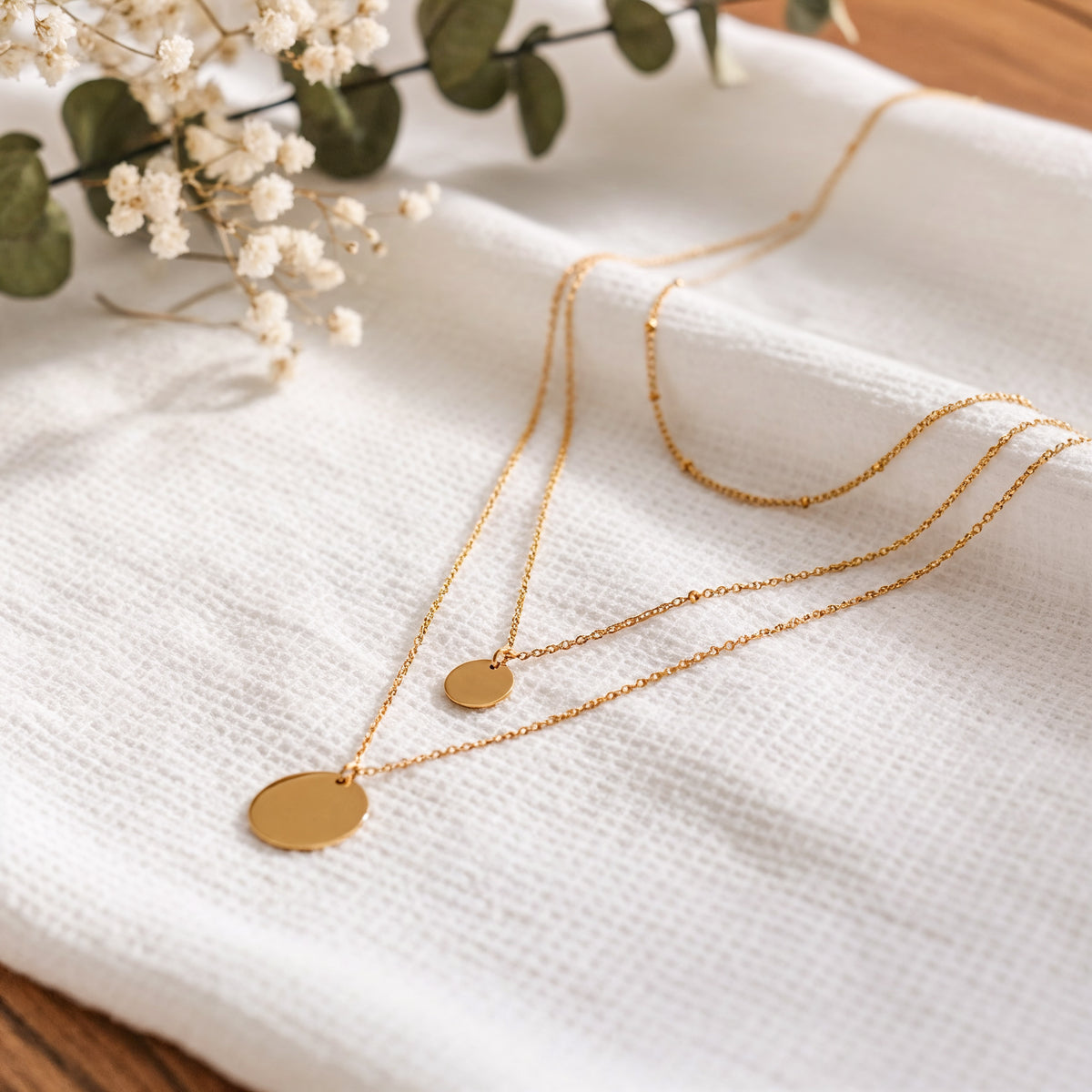Dreilagige Halskette mit runden Plättchen aus Edelstahl 18 K vergoldet, liegt auf weißem Leinentuch mit Trockenblumen – Elysian Layers Kette | Mercy’s Jewelry