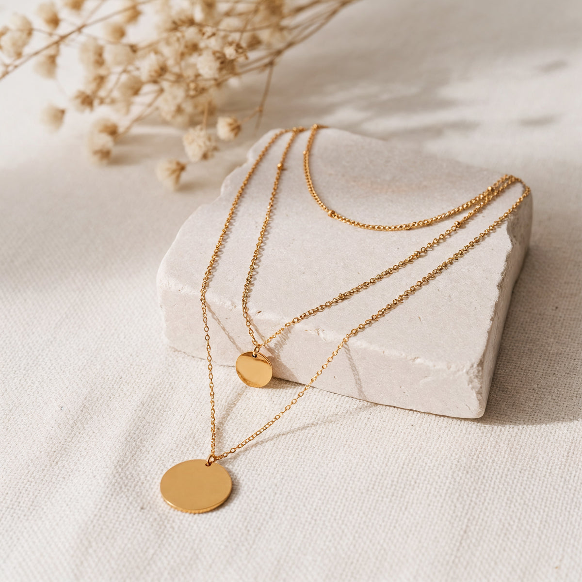 Dreilagige Halskette mit runden Plättchen aus Edelstahl 18 K vergoldet, liegt auf heller Steinplatte mit Trockenblumen – Elysian Layers Kette | Mercy’s Jewelry
