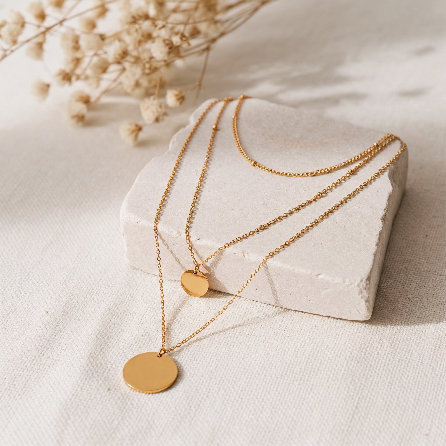 Dreilagige Halskette mit runden Plättchen aus Edelstahl 18 K vergoldet, liegt auf heller Steinplatte mit Trockenblumen – Elysian Layers Kette | Mercy’s Jewelry