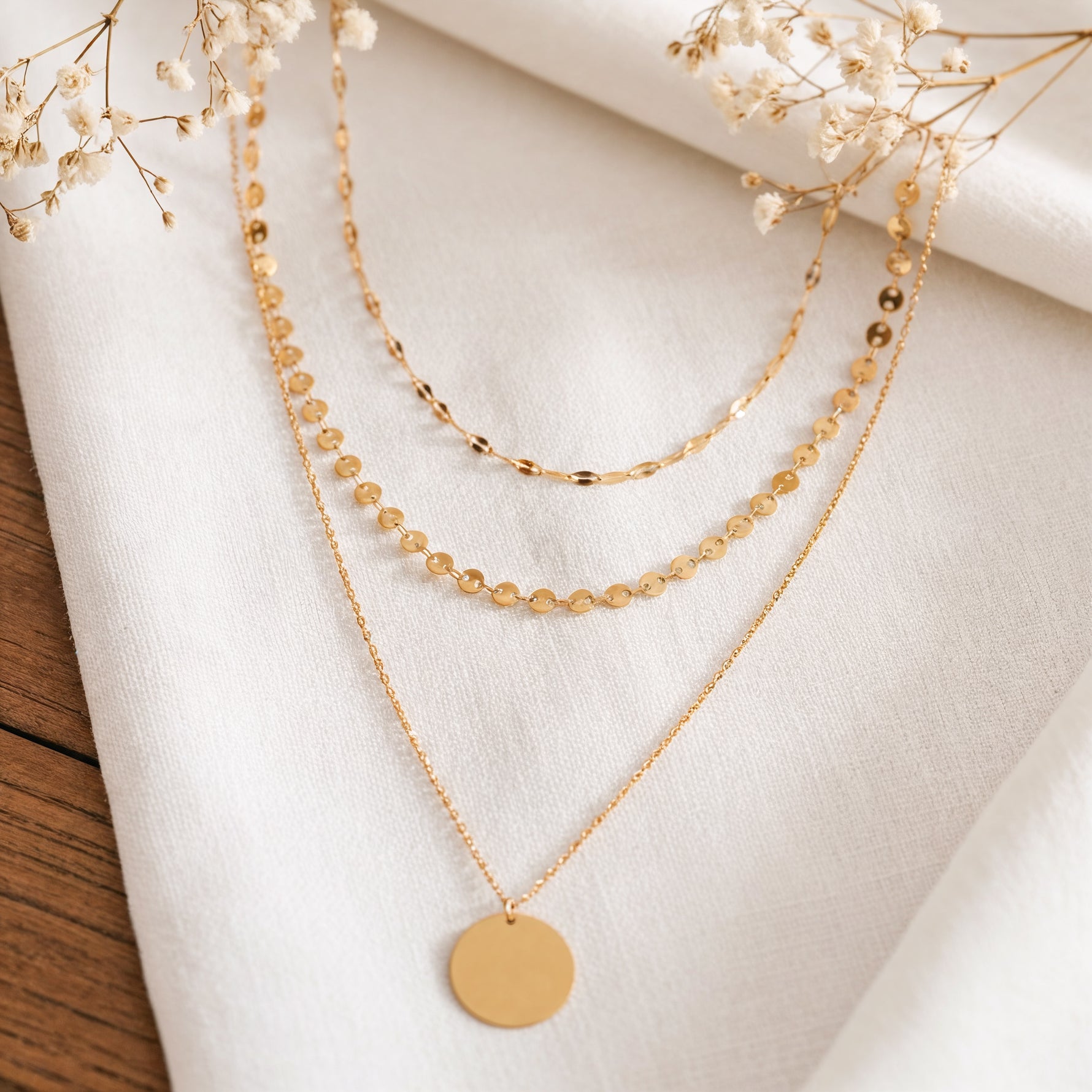 Dreilagige Halskette mit Plättchen-, Kugel- und Gliederdesign aus Edelstahl 18 K vergoldet, liegt auf weißem Leinentuch mit Trockenblumen – Aurora Layers Kette | Mercy’s Jewelry