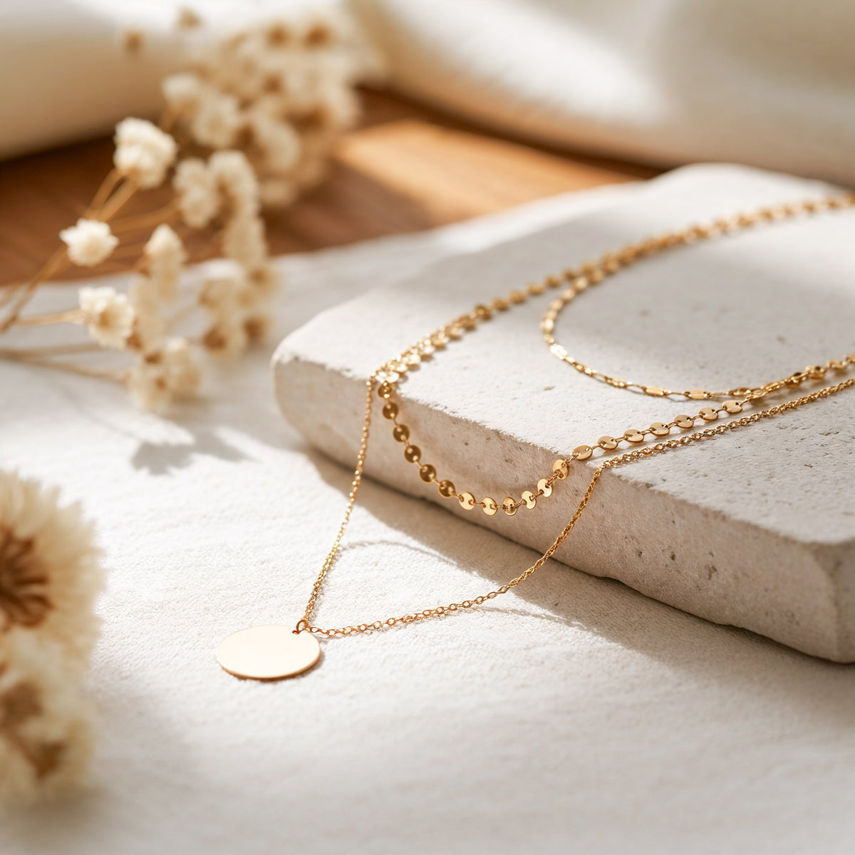 Dreilagige Halskette mit Plättchen-, Kugel- und Gliederdesign aus Edelstahl 18 K vergoldet, liegt auf heller Steinplatte mit Trockenblumen im Sonnenlicht – Aurora Layers Kette | Mercy’s Jewelry