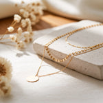 Dreilagige Halskette mit Plättchen-, Kugel- und Gliederdesign aus Edelstahl 18 K vergoldet, liegt auf heller Steinplatte mit Trockenblumen im Sonnenlicht – Aurora Layers Kette | Mercy’s Jewelry