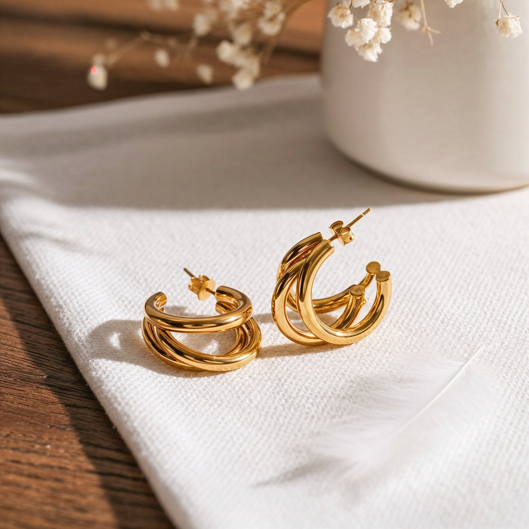 Dreireihige Creolen aus Edelstahl 18 K vergoldet mit glänzendem Finish, liegen auf hellem Stoff mit Feder und Trockenblumen – Taviel Creolen | Mercy’s Jewelry