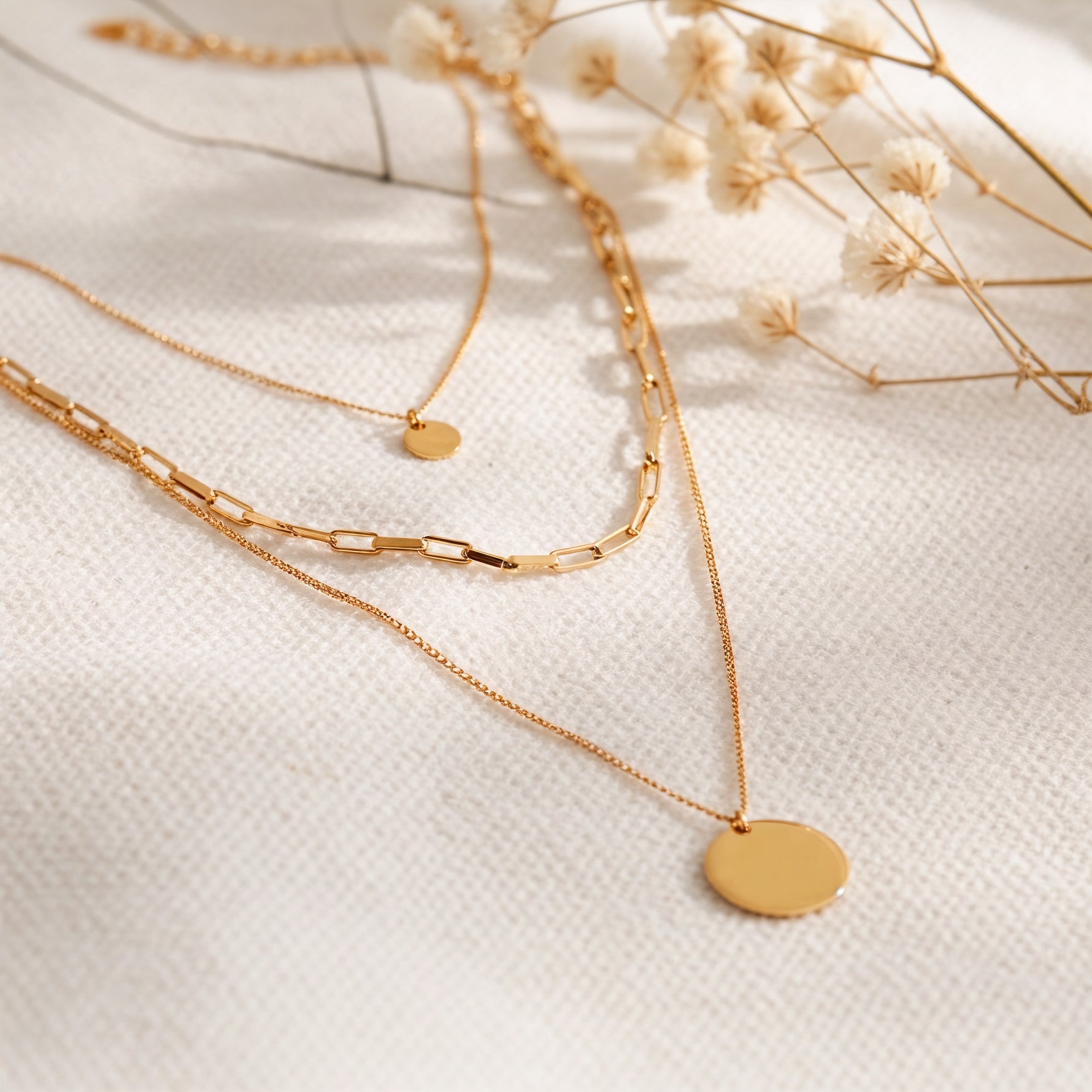 Dreireihige Gliederkette mit rundem Anhänger aus Edelstahl 18 K vergoldet, liegt auf hellem Stoff mit Trockenblumen im warmen Sonnenlicht – Sienna Layers Kette | Mercy’s Jewelry