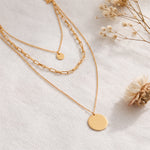 Dreireihige Gliederkette mit rundem Anhänger aus Edelstahl 18 K vergoldet, liegt auf hellem Stoff mit Trockenblumen im Tageslicht – Sienna Layers Kette | Mercy’s Jewelry