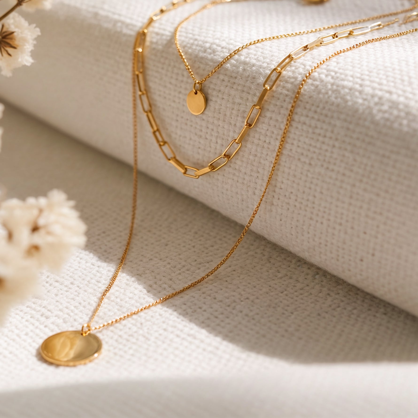 Dreireihige Gliederkette mit rundem Anhänger aus Edelstahl 18 K vergoldet, liegt auf hellem Stoff mit sanftem Licht und Trockenblumen – Sienna Layers Kette | Mercy’s Jewelry