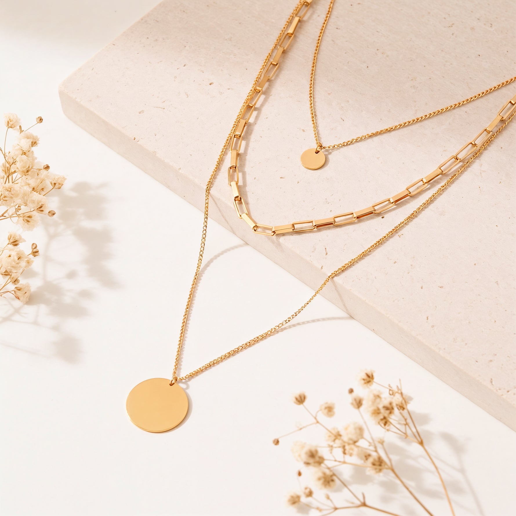 Dreireihige Gliederkette mit rundem Anhänger aus Edelstahl 18 K vergoldet, liegt auf heller Steinplatte mit Trockenblumen im Sonnenlicht – Sienna Layers Kette | Mercy’s Jewelry