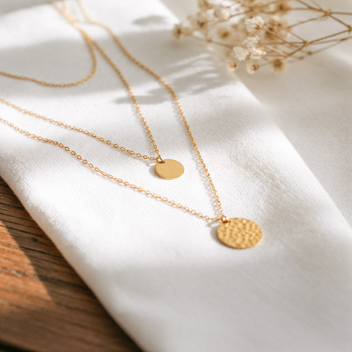 Dreireihige Halskette mit glattem und gehämmertem Anhänger aus Edelstahl 18 K vergoldet, liegt auf weißem Leinentuch mit Holzuntergrund im warmen Tageslicht – Vela Layers Kette | Mercy’s Jewelry
