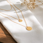 Dreireihige Halskette mit glattem und gehämmertem Anhänger aus Edelstahl 18 K vergoldet, liegt auf weißem Leinentuch mit Holzuntergrund im warmen Tageslicht – Vela Layers Kette | Mercy’s Jewelry