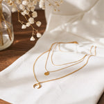 Dreireihige Halskette mit rundem und Halbmond-Anhänger aus Edelstahl 18 K vergoldet, liegt auf weißem Stoff mit Holzhintergrund und getrockneten Blumen – Noenya Layers Halskette | Mercy’s Jewelry