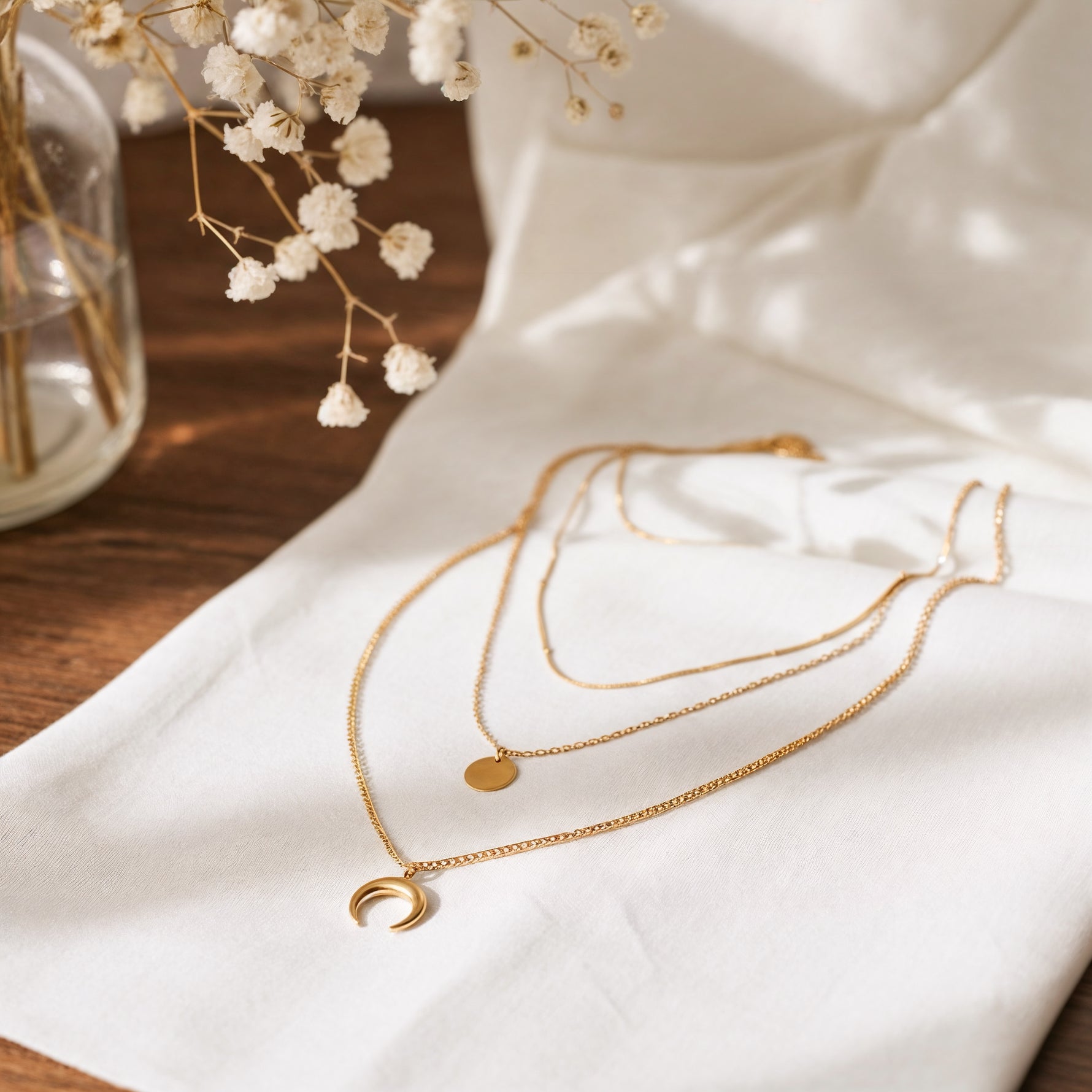 Dreireihige Halskette mit rundem und Halbmond-Anhänger aus Edelstahl 18 K vergoldet, liegt auf weißem Stoff mit Holzhintergrund und getrockneten Blumen – Noenya Layers Halskette | Mercy’s Jewelry