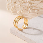 Dreireihiger Ring aus Edelstahl 18 K vergoldet, mit geflochtener Struktur am Rand auf heller Steinplattform mit Trockenblumen im Hintergrund – Nirella Ring | Mercy’s Jewelry