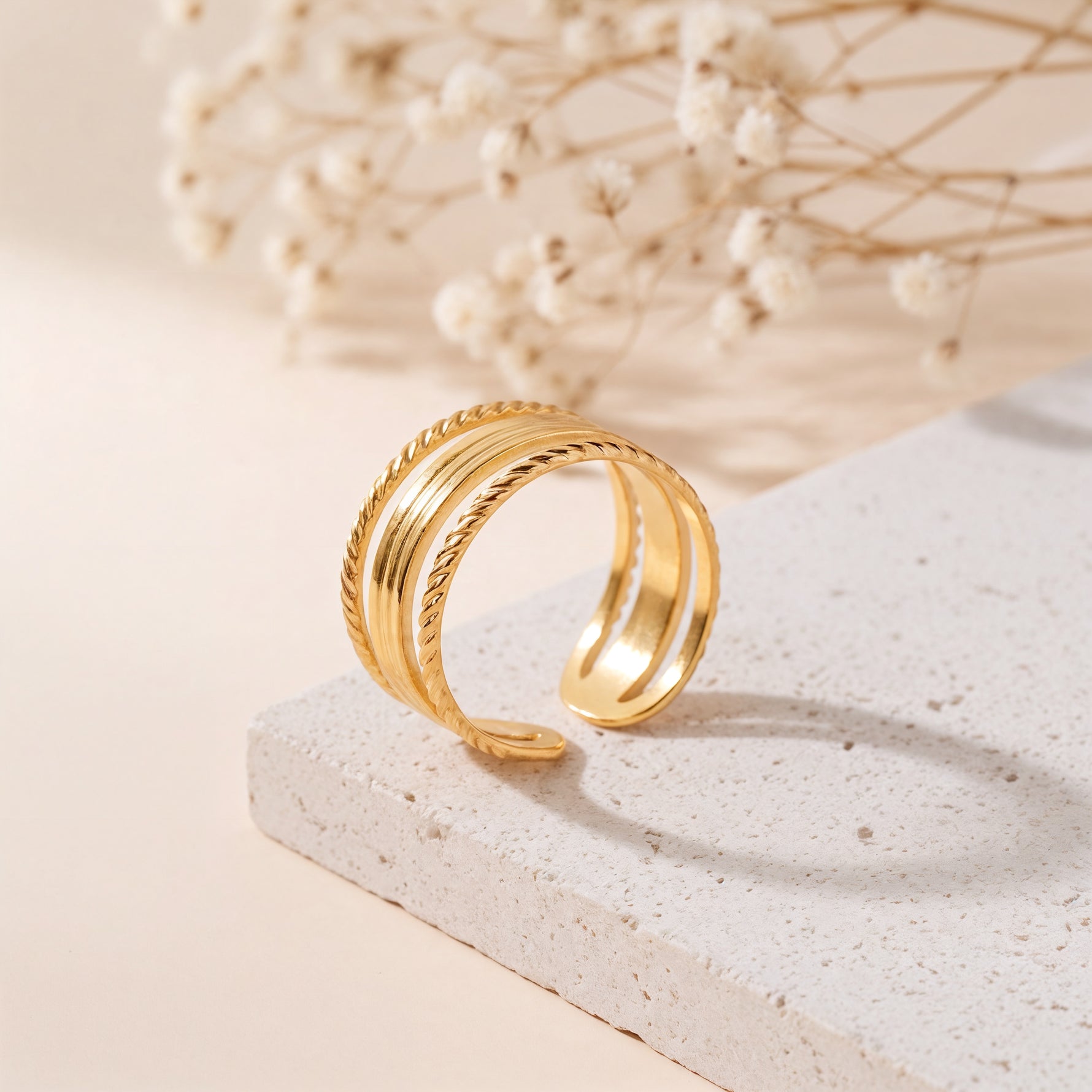 Dreireihiger Ring aus Edelstahl 18 K vergoldet, mit geflochtener Struktur am Rand auf heller Steinplattform mit Trockenblumen im Hintergrund – Nirella Ring | Mercy’s Jewelry