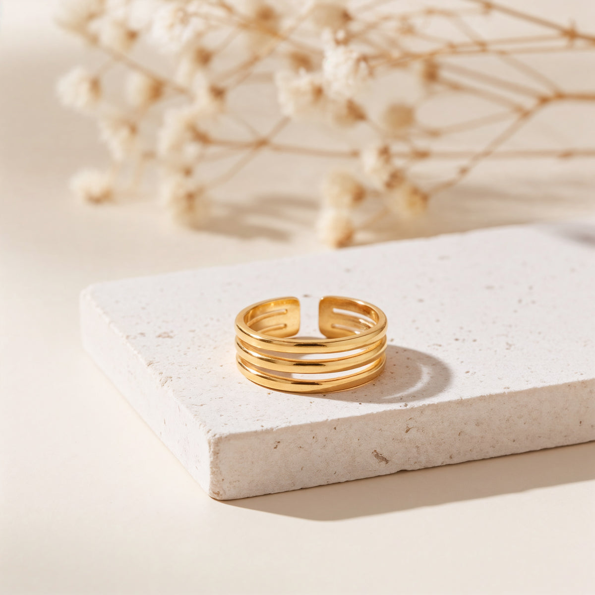 Dreireihiger Ring aus Edelstahl 18 K vergoldet, minimalistisches Design auf heller Steinplattform mit Trockenblumen im Hintergrund – Kelana Ring | Mercy’s Jewelry