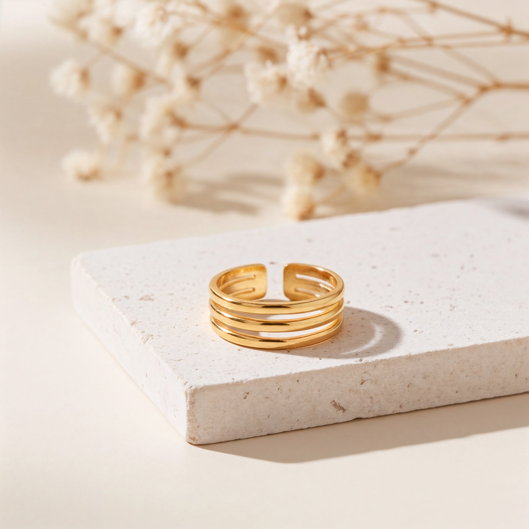 Dreireihiger Ring aus Edelstahl 18 K vergoldet, minimalistisches Design auf heller Steinplattform mit Trockenblumen im Hintergrund – Kelana Ring | Mercy’s Jewelry