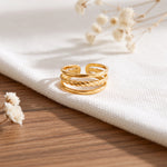 Dreireihiger Ring aus Edelstahl 18 K vergoldet, offen und verstellbar, mit strukturierter und gedrehter Oberfläche auf weißem Stofftuch mit Holzuntergrund – Alirae Ring | Mercy’s Jewelry