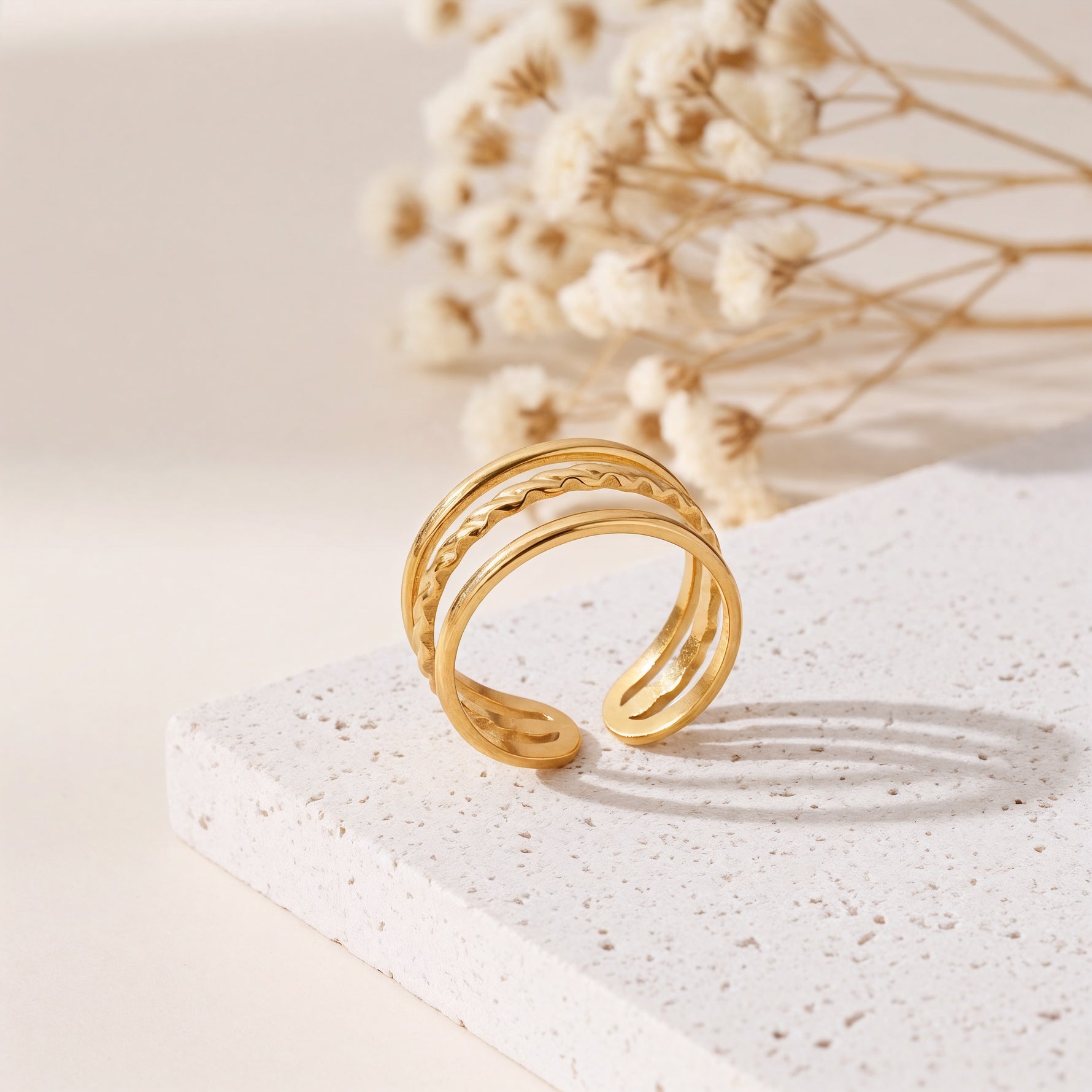 Dreireihiger Ring aus Edelstahl 18 K vergoldet, offen und verstellbar, mit strukturierter Oberfläche auf heller Steinplattform und Trockenblumen im Hintergrund – Alirae Ring | Mercy’s Jewelry