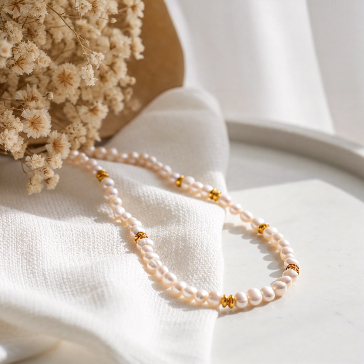 Feine Perlenkette mit goldenen Zwischenelementen aus Edelstahl 18 K vergoldet, liegt auf hellem Stoff mit Trockenblumen und runder Steinplatte – Orielle Perlenkette | Mercy’s Jewelry