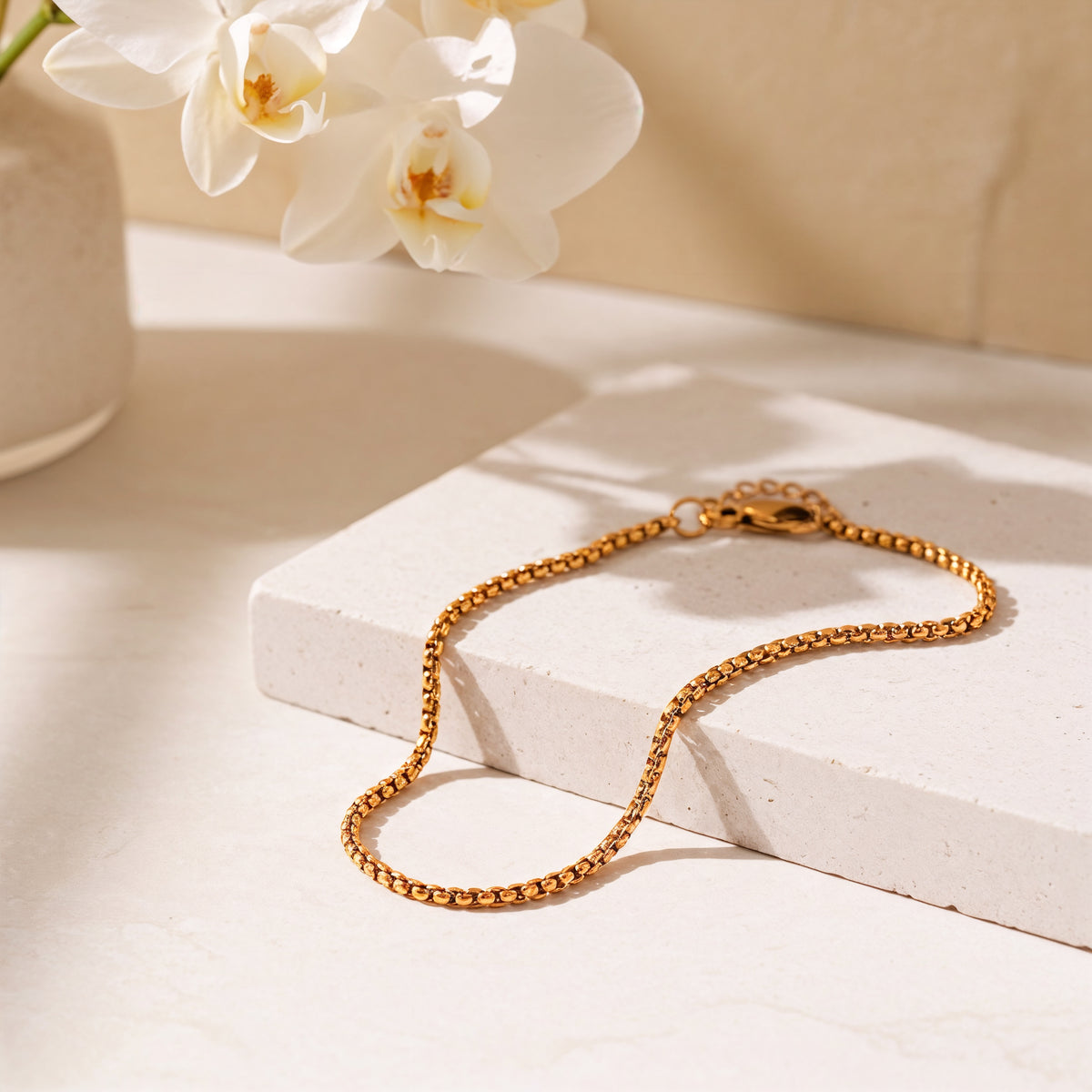 Feines Armband aus Edelstahl 18 K vergoldet mit zarter Gliederstruktur, liegend auf hellem Steinuntergrund im Sonnenlicht neben weißen Orchideen – Amelira Armband | Mercy’s Jewelry