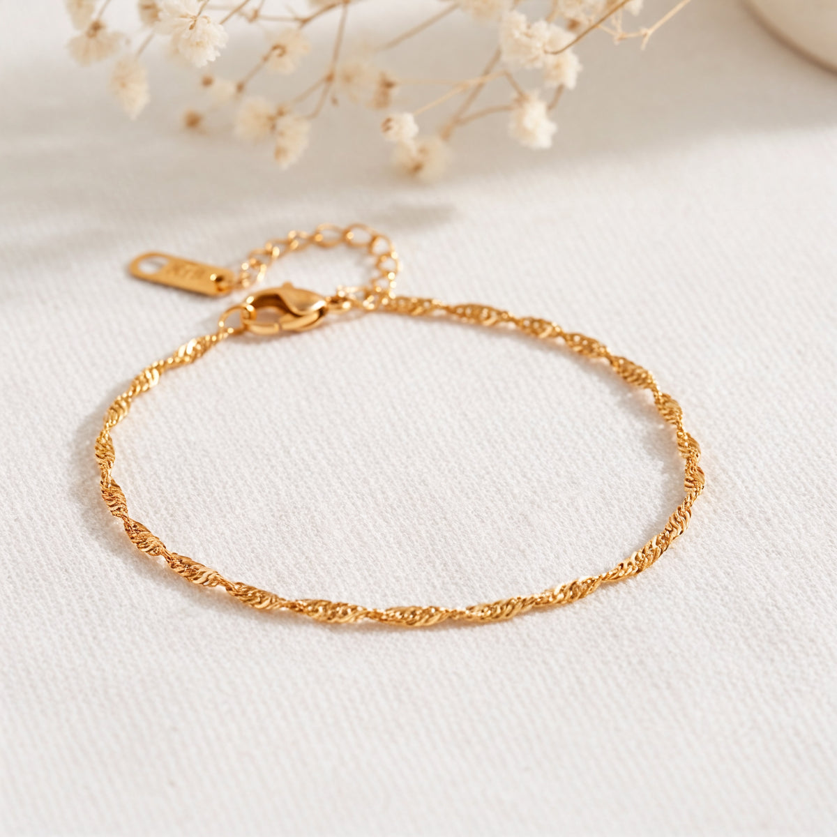 Feines Rope-Armband aus Edelstahl 18 K vergoldet mit gedrehter Struktur, liegend auf hellem Stoff mit Trockenblumen – Fayara Armband | Mercy’s Jewelry