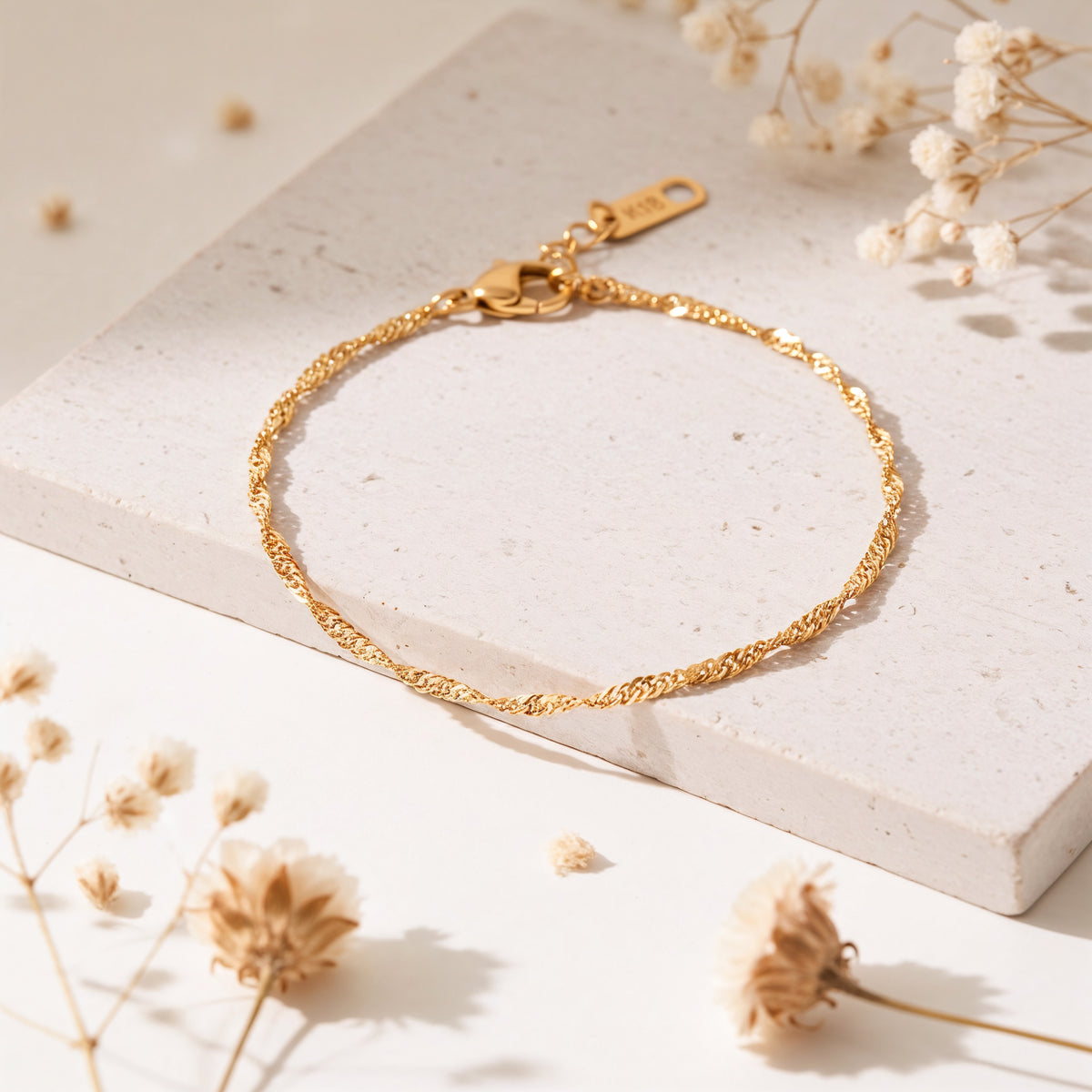 Feines Rope-Armband aus Edelstahl 18 K vergoldet mit gedrehter Struktur, elegant auf heller Steinplatte mit Trockenblumen arrangiert – Fayara Armband | Mercy’s Jewelry