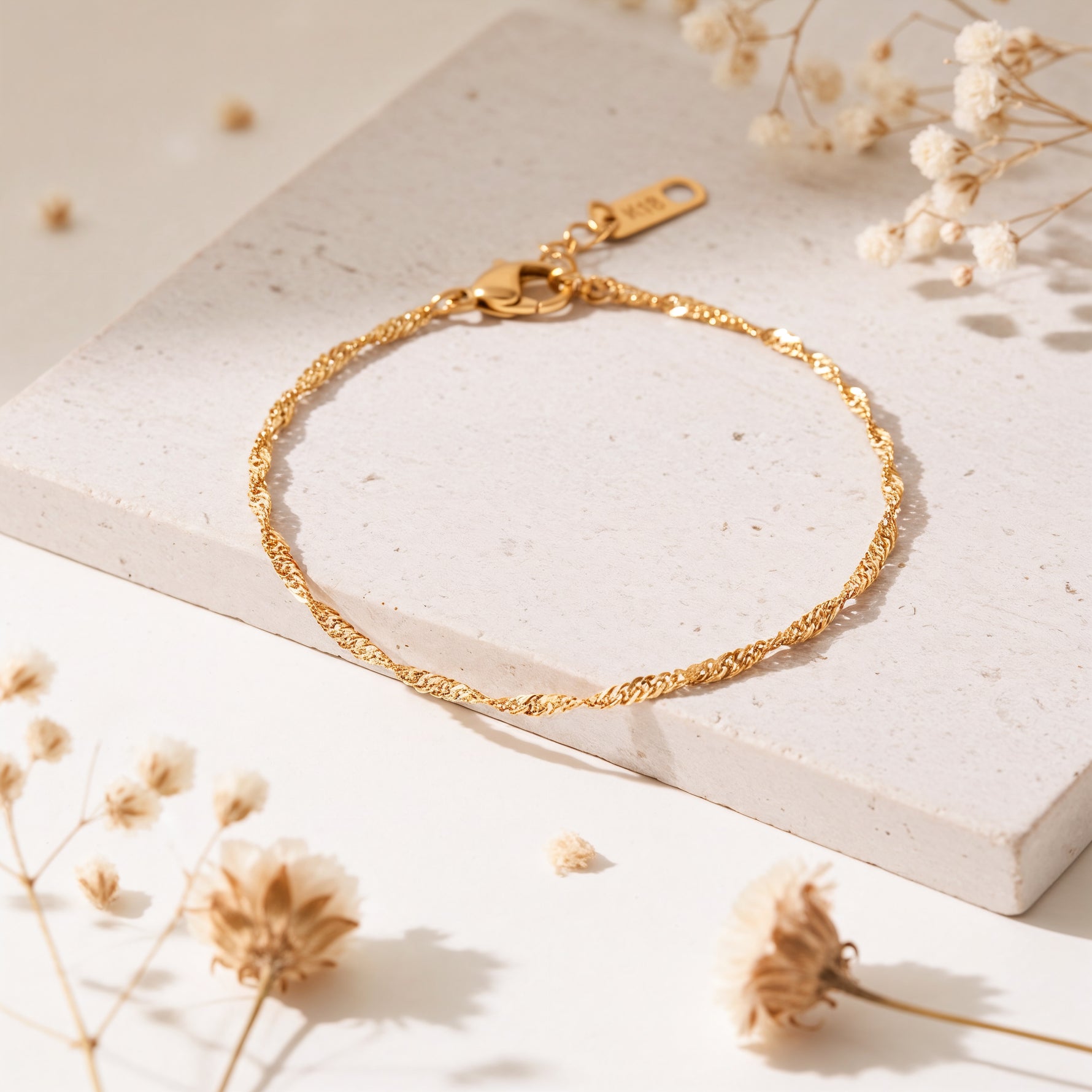 Feines Rope-Armband aus Edelstahl 18 K vergoldet mit gedrehter Struktur, elegant auf heller Steinplatte mit Trockenblumen arrangiert – Fayara Armband | Mercy’s Jewelry