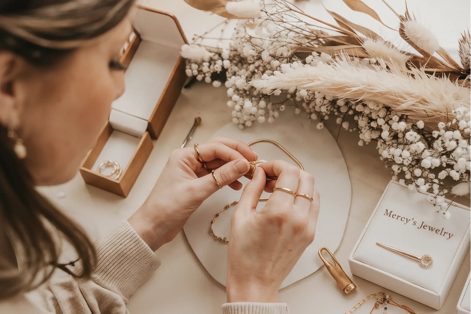 Frau legt eine goldene Halskette an, umgeben von weiteren Schmuckstücken, einer Mercy’s Jewelry Box und trockenen Pampas- und Baby’s-Breath-Blumen in warmem Licht.