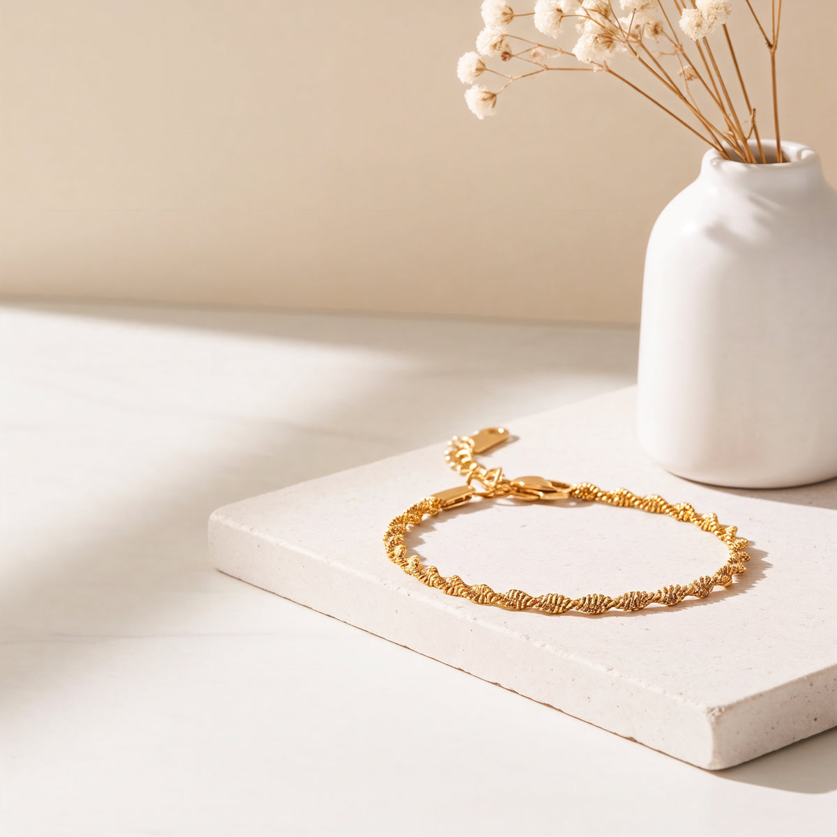 Feines, gedrehtes Armband aus Edelstahl 18 K vergoldet, auf heller Steinplatte mit Vase und Trockenblumen im Sonnenlicht – Solira Armband | Mercy’s Jewelry