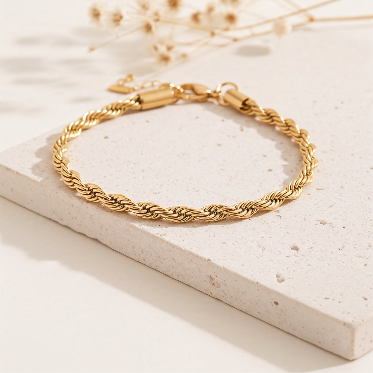 Gedrehtes Seilarmband aus Edelstahl 18 K vergoldet mit polierter Oberfläche, stilvoll auf heller Steinplatte mit Trockenblumen – Tharien Armband | Mercy’s Jewelry