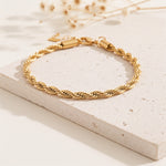 Gedrehtes Seilarmband aus Edelstahl 18 K vergoldet mit polierter Oberfläche, stilvoll auf heller Steinplatte mit Trockenblumen – Tharien Armband | Mercy’s Jewelry