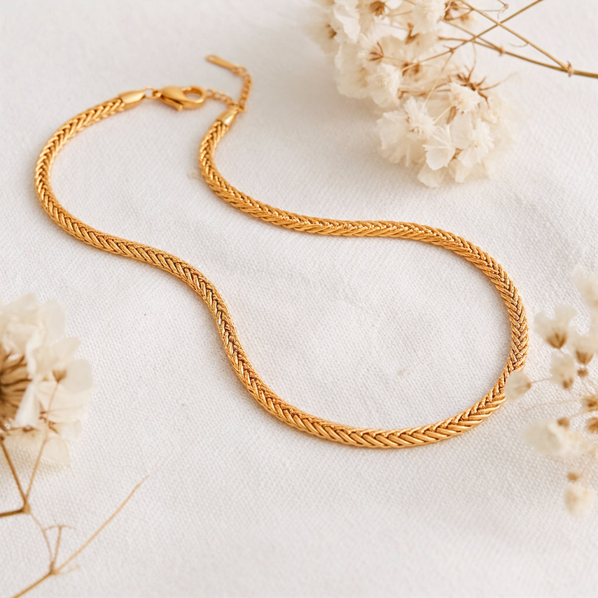 Geflochtene Halskette aus Edelstahl 18 K vergoldet, liegt auf weißem Leinentuch mit Trockenblumen im warmen Tageslicht – Kaelwyn Kette | Mercy’s Jewelry