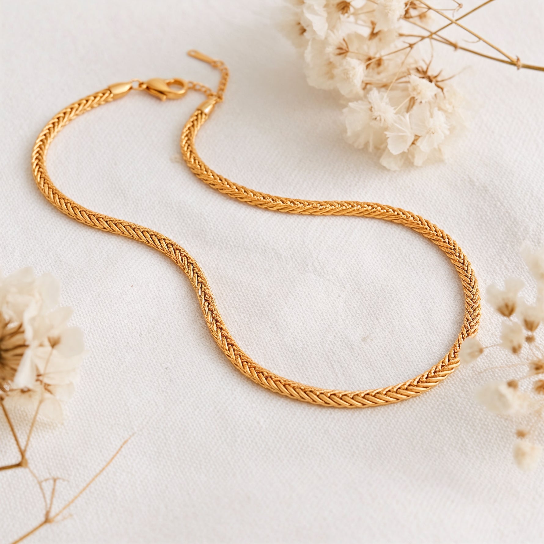 Geflochtene Halskette aus Edelstahl 18 K vergoldet, liegt auf weißem Leinentuch mit Trockenblumen im warmen Tageslicht – Kaelwyn Kette | Mercy’s Jewelry