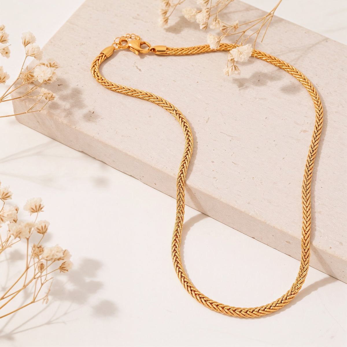 Geflochtene Halskette aus Edelstahl 18 K vergoldet, liegt auf heller Steinplatte mit Trockenblumen im Sonnenlicht – Kaelwyn Kette | Mercy’s Jewelry