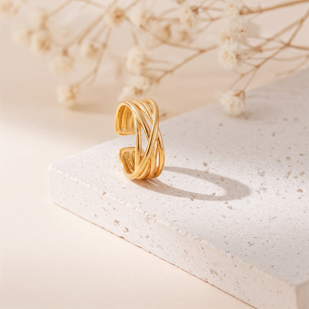 Gekreuzter Ring aus Edelstahl 18 K vergoldet, offen und verstellbar, mit elegant geschwungenem Design auf heller Steinplattform mit Trockenblumen im Hintergrund – Elanira Ring | Mercy’s Jewelry