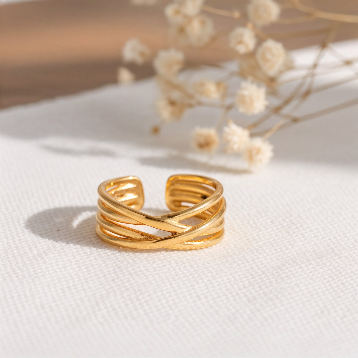 Gekreuzter Ring aus Edelstahl 18 K vergoldet, offen und verstellbar, mit elegant geschwungenem Design auf hellem Stoff mit Trockenblumen im Hintergrund – Elanira Ring | Mercy’s Jewelry