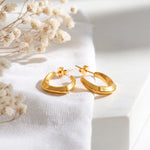 Geometrische Creolen mit kantigem Design aus Edelstahl 18 K vergoldet, liegen auf hellem Stoff mit Trockenblumen im Hintergrund – Virel Creolen | Mercy’s Jewelry